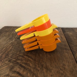 May include: A set of four yellow and orange plastic measuring cups. The cups are stacked on top of each other, with the largest cup on the bottom and the smallest cup on top.