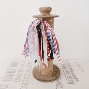 May include: A wooden spool with a variety of red, white, and blue ribbons tied around it. The spool is sitting on a white surface with text printed on it.