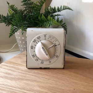 May include: A vintage white Westclox kitchen timer with a cream-colored dial and black numerals. The timer has a knob and a scale marked from 0 to 60. The text "TURN PAST STAR THEN SET TIME" is visible. The timer sits on a wooden surface.