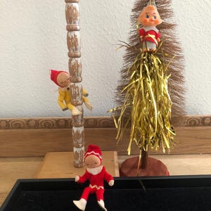 May include: A collection of vintage Christmas decorations featuring small elf figurines. One elf is climbing a wooden post, another is atop a miniature tree with gold tinsel, and a third is seated on a black tray. The elves wear red and yellow outfits.