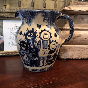 May include: A ceramic pitcher with a cream-colored base and blue painted details. The pitcher features a whimsical design of smiling figures and hearts. The handle and rim are also painted blue, creating a cohesive and charming aesthetic.