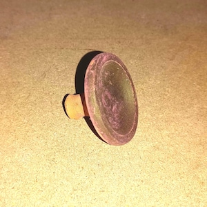 Puede incluir: Un pequeño tapón de botella redondo con un tallo corto y cilíndrico. El tapón tiene un tono rosado-púrpura en la superficie superior y un tallo amarillo dorado. El artículo probablemente está hecho de vidrio o un material similar.