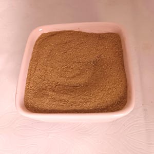 May include: A square, light pink ceramic bowl filled with a finely ground, medium-brown powder. The powder has a smooth, textured surface that fills the entire interior of the bowl, creating a uniform appearance with a consistent earthy color.
