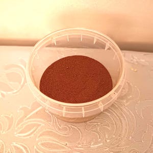 May include: A clear plastic container holding a reddish-brown granular material. The container is round with a lid. The substance is evenly distributed inside. The container sits on a patterned surface.
