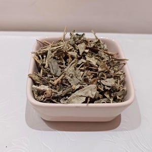 May include: A square, light pink bowl filled with dried green herbs. The herbs have a variety of leaf shapes and textures, with some stems visible. The bowl is sitting on a white surface.