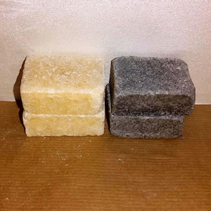 Pode incluir: Duas barras de sabão retangulares estão posicionadas lado a lado. A barra à esquerda é de uma cor amarelo-pálido translúcida com uma superfície texturizada. A barra à direita é de cor cinza-carvão escuro com uma textura granulada e porosa.