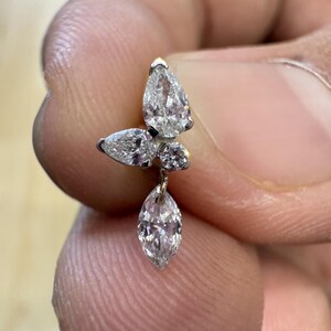 Może przedstawiać: Zbliżenie kolczyka z diamentami o motywie motyla. Kolczyk ma pięć przezroczystych, błyszczących diamentów w kształcie gruszki i okrągłym, osadzonych w srebrnym metalu. Kolczyk jest trzymany między palcami.