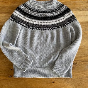 Puede incluir: Un suéter gris claro con cuello alto y acanalado. El canesú presenta un patrón geométrico en negro, blanco y gris. El suéter tiene mangas largas y dobladillo y puños acanalados. La prenda está hecha de un material de punto suave.