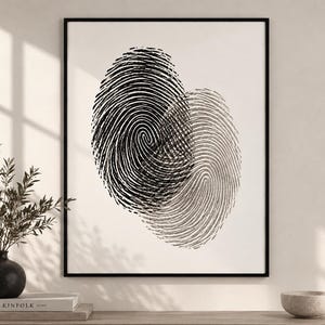 May include: A framed wall art print featuring two overlapping fingerprint patterns in black and dark gray ink on a white background. The artwork is enclosed in a thin black frame, displaying intricate, swirling ridge details that form a central focal point.