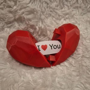 May include: A red, geometric heart-shaped box, split in two, with a white card inside. The card reads "I ❤️ You" in black text with a red heart symbol. The box is on a fluffy, white surface.