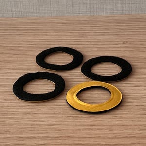 Può includere: Quattro anelli adesivi in feltro nero con retro giallo sono disposti su una superficie di legno. Gli anelli in feltro hanno una forma circolare con un foro al centro. Il retro adesivo è visibile su uno degli anelli.