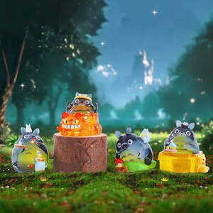 May include: A collection of translucent figurines of the character Totoro from the animated film My Neighbor Totoro. The figurines are in various poses and colors, set against a forest backdrop with a wooden stump.