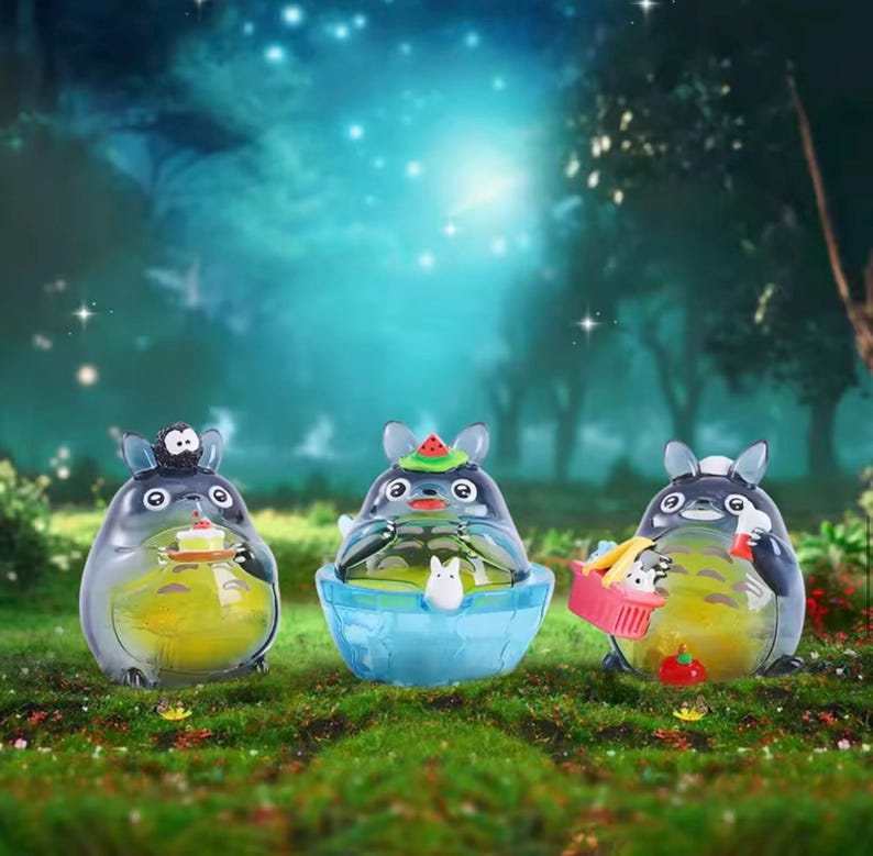 May include: Three whimsical Totoro figurines. One holds a cake, another sits in a blue bowl, and the third carries a picnic basket. Each figure has a gray body, yellow belly, and black eyes. The background is a blurred forest scene.