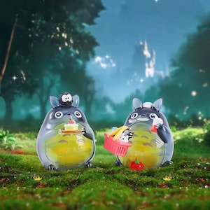 May include: Two translucent Totoro figures with yellow liquid interiors stand on green grass. One holds a cake, the other a picnic basket with a small animal and a red apple. The background features a forest and a starry sky.