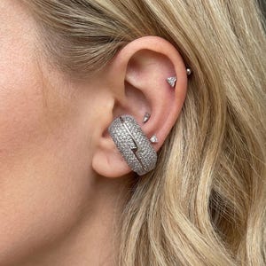 May include: A silver ear cuff with a lightning bolt design, encrusted with small, sparkling stones. The cuff is worn on the ear, alongside several small, silver stud earrings. The ear is framed by blonde hair.