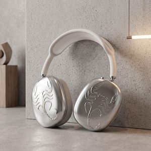 May include: Over-ear headphones featuring a white headband and metallic silver ear cups. Each ear cup is embossed with a detailed scorpion design. The headphones have a smooth, rounded aesthetic with visible mesh padding on the underside of the headband.