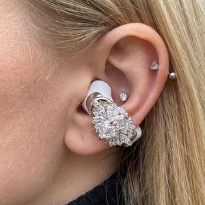 Op de afbeelding: Witte oortelefoons met een zilveren en met diamanten bezet decoratief element. De oortelefoon heeft een grote ovaal geslepen diamant omringd door kleinere diamanten. De afbeelding toont de oortelefoon in het oor van een persoon, samen met meerdere piercings.