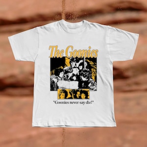 May include: White t-shirt featuring a graphic from the movie "The Goonies." The design includes the film's title in yellow and a black and white image of the cast. The phrase "Goonies never say die!" is printed below the image.