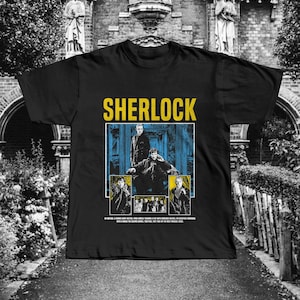 May include: Black t-shirt featuring the word "SHERLOCK" in yellow block letters above a graphic of the show's characters. The graphic is in blue, yellow, and black. Below the graphic is a line of text.