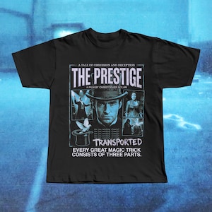 Könnte beinhalten: Schwarzes T-Shirt mit einem Filmplakat-Design für "The Prestige" von Christopher Nolan. Das Design zeigt den Filmtitel, Bilder und den Text "TRANSPORTED". Das Design ist in Blau- und Weißtönen gehalten. Das T-Shirt hat einen Rundhalsausschnitt.