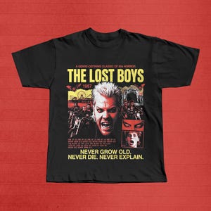 May include: Black t-shirt featuring a movie poster design for "The Lost Boys" from 1987. The design includes the movie title, images of characters, and the text "Never Grow Old. Never Die. Never Explain."