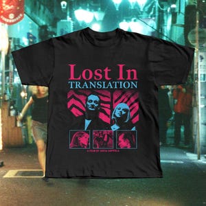 以下が含まれることがあります： ピンクと青の文字で「Lost In Translation」という映画タイトルがプリントされた黒いTシャツ。2人の人物と3つの小さな画像が描かれています。「A Film by Sofia Coppola」というテキストも含まれています。