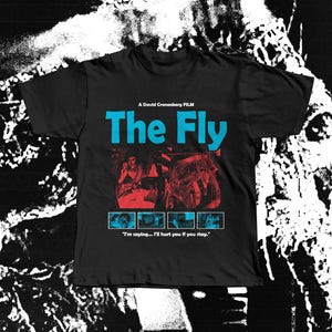 Puede incluir: Camiseta negra con el título de la película "The Fly" en azul. La camiseta muestra un gráfico de la película y la cita: "I'm saying... I'll hurt you if you stay." El texto "A David Cronenberg FILM" está encima del título.