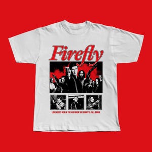 Puede incluir: Camiseta blanca con un diseño gráfico de la serie de televisión Firefly. El diseño incluye el título del programa en escritura roja sobre una imagen en blanco y negro del elenco con armas. El texto "LOVE KEEPS HER IN THE AIR WHEN SHE OUGHTTA FALL DOWN" está debajo.