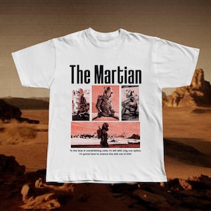 May include: White t-shirt featuring the text "The Martian" above four images of a space-suited figure. Below the images is the quote, "In the face of overwhelming odds, I'm left with only one option. I'm gonna have to science the sh*t out of this."