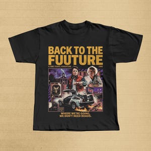Könnte beinhalten: Schwarzes T-Shirt mit einer Grafik aus dem Film "Zurück in die Zukunft". Das Design zeigt den Text "BACK TO THE FUUTURE" in Gold, Bilder der DeLorean-Zeitmaschine und Charaktere. Der Text "WHERE WE'RE GOING, WE DON'T NEED ROADS" ist ebenfalls enthalten.
