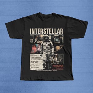 May include: Black t-shirt featuring the movie title "Interstellar" in white block letters. The shirt has a graphic of an astronaut and scenes from the film. Text includes "Mission: Save Humanity" and "Directed by Christopher Nolan."
