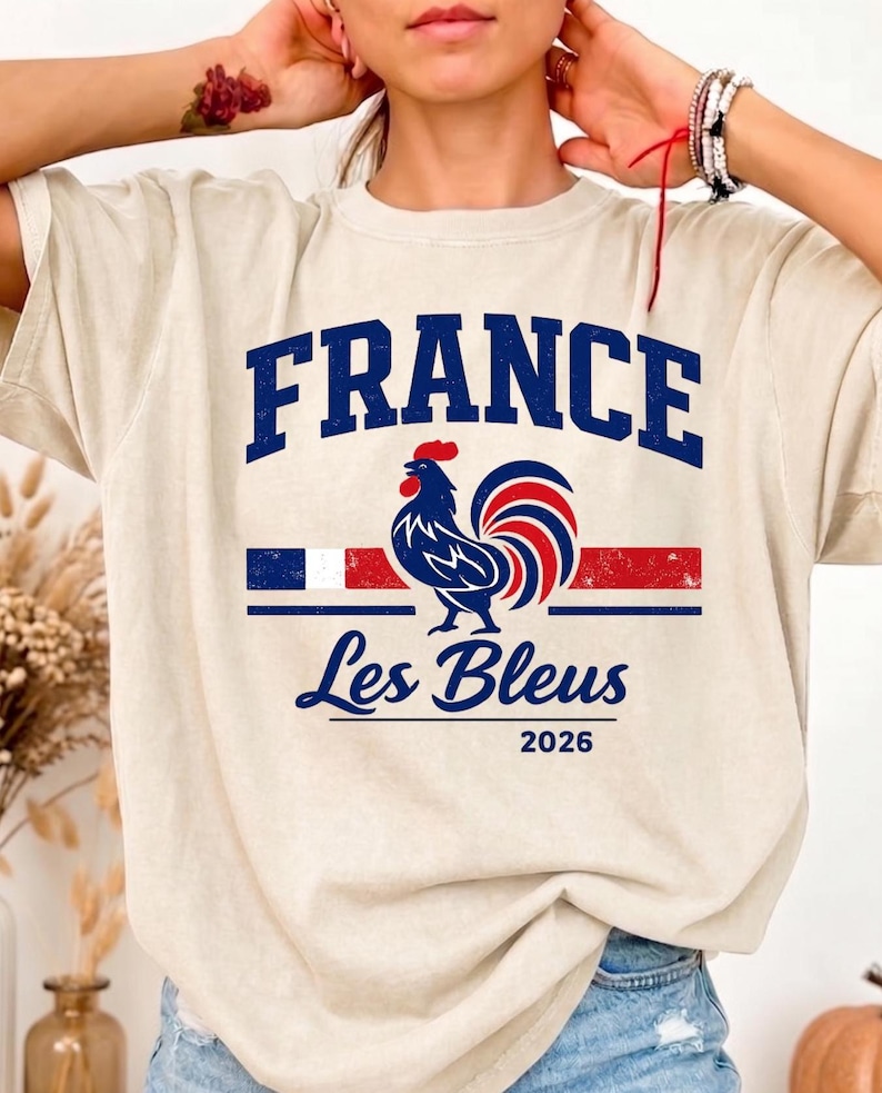 May include: A beige short-sleeve t-shirt featuring a graphic with the text FRANCE in blue, a blue rooster illustration, a horizontal tricolor stripe, the text Les Bleus in blue script, and the number 2026 printed in blue on the front.