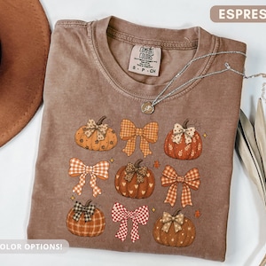 Puede incluir: Una camiseta color espresso con un estampado gráfico de cinco calabazas y cuatro lazos en varios patrones, incluyendo cuadros, vichy y estampado de leopardo. La camiseta tiene cuello redondo y una etiqueta de la marca Comfort Colors.