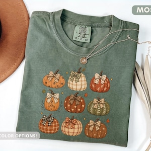 Puede incluir: Una camiseta de color verde musgo con un estampado gráfico de nueve calabazas, cada una decorada con un lazo estampado único. Las calabazas varían en color y textura, incluyendo tonos naranja, crema y verde. La camiseta tiene cuello redondo y etiqueta.