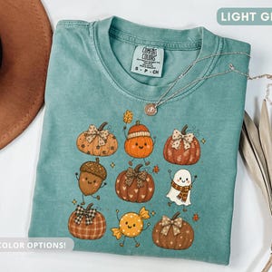 Puede incluir: Una camiseta verde claro con un estampado gráfico de varios personajes otoñales, incluyendo calabazas, una bellota y un fantasma. La camiseta tiene cuello redondo y una etiqueta de la marca Comfort Colors. Un collar plateado descansa sobre ella.