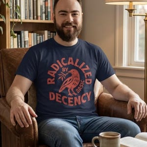 Pode incluir: Camiseta azul marinho com o texto "RADICALIZED BY BASIC DECENCY" em um design circular com um pássaro vermelho segurando um ramo de oliveira. A pessoa está usando jeans azuis e sentada em uma poltrona de couro marrom.