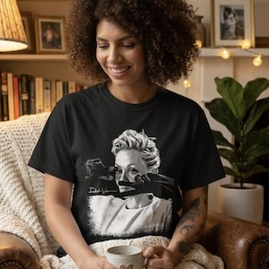 May include: A black short-sleeved t-shirt featuring a white graphic print of a person with short hair wearing dark gloves over their eyes. The print includes a signature and a rectangular border. The shirt is made of a solid black fabric.