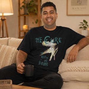 May include: Black t-shirt featuring the band "The Cure" with the album title "The Head on the Door" in teal and white. The shirt has a graphic design of a cloud-like image. The person is sitting on a beige couch and holding a dark gray mug.
