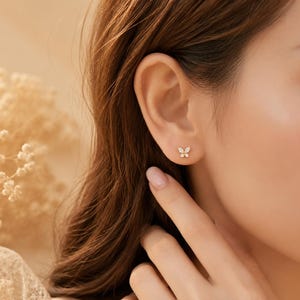 May include: A small gold stud earring shaped like a butterfly, featuring four clear, marquise-cut stones arranged to form the wings. The earring is positioned on the earlobe, showcasing a delicate metal frame and a polished, reflective finish.