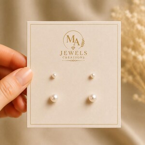 May include: Two pairs of white spherical pearl stud earrings mounted on a rectangular cream-colored display card. The card features the gold-toned logo and text MA JEWELS CREATIONS centered at the top above the four individual pearl studs.