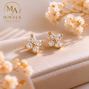 Puede incluir: Un par de pendientes de botón dorados con forma de flor con gemas claras y brillantes. Los pendientes se muestran sobre una superficie beige suave. El logotipo "MA JEWELS CREATIONS" es visible.