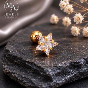 Pode incluir: Um brinco dourado em forma de estrela com uma pedra central transparente e facetada. O brinco tem uma parte traseira em forma de bola e está colocado sobre uma superfície escura e texturizada. A imagem também apresenta o texto "MA JEWELS CREATIONS".