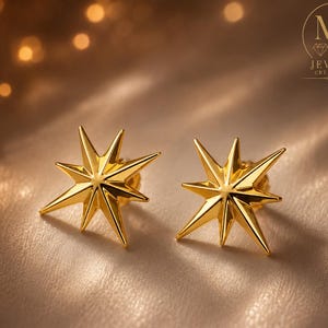 May include: A pair of gold-colored starburst stud earrings. Each earring features eight pointed rays emanating from a central point. The earrings are displayed on a textured, neutral-toned surface. The logo "MA Jewels Creations" is visible.