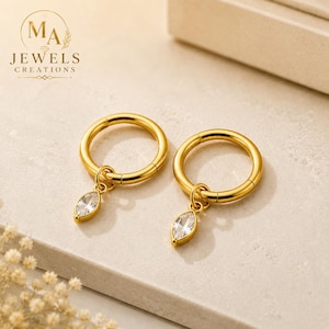 May include: A pair of gold-toned hoop earrings, each featuring a single marquise-cut clear gemstone charm suspended from the circular band. The jewelry displays a polished metallic finish and includes the logo text MA JEWELS CREATIONS.