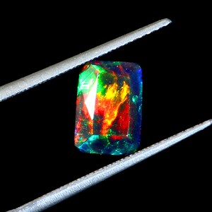 May include: A rectangular black opal gemstone with vibrant internal colors of red, yellow, green, and blue. The gemstone is held by silver tweezers against a black background. The opal has a polished, faceted surface.