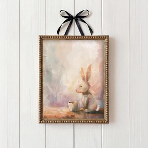 May include: A framed art print depicting a white rabbit with closed eyes, beside a small cup. The artwork uses a soft pastel colour palette and is framed in a gold-coloured frame, with a black ribbon for hanging. The frame measures approximately 25 cm x 25 cm.