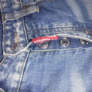 May include: Close-up of blue denim jeans with visible stitching and metal button closures. A red tag with white text is attached to the jeans. The jeans have a classic five-pocket design.