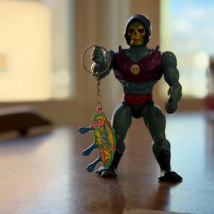 May include: A Skeletor action figure with a skull-like face, wearing a purple chest piece and red trunks. He holds a colorful, abstract charm. The figure is gray and black, with a yellow skull and black boots.