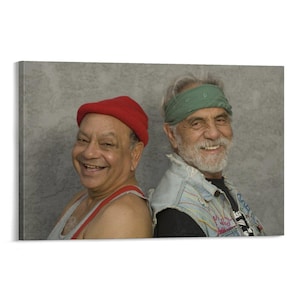 May include: A canvas print featuring two smiling men leaning back-to-back. One man wears a red knit cap and a gray tank top with red suspenders. The other man wears a green bandana and a denim vest over a black shirt.