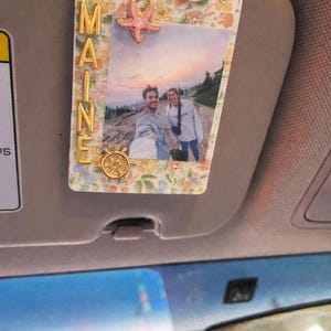 May include: A decorative car visor photo holder with a floral pattern and the word "MAINE" in gold letters. The holder features a photo of two people and is adorned with a pink starfish, a pink whale, and a gold ship's wheel.